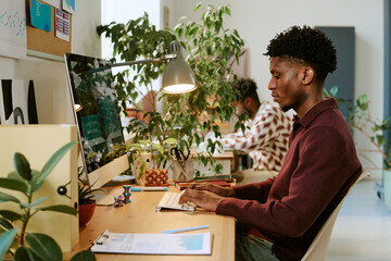 Young adult Black man working at computer in modern office, typing on keyboard with focused expression, surrounded by plants and documents, another working in background