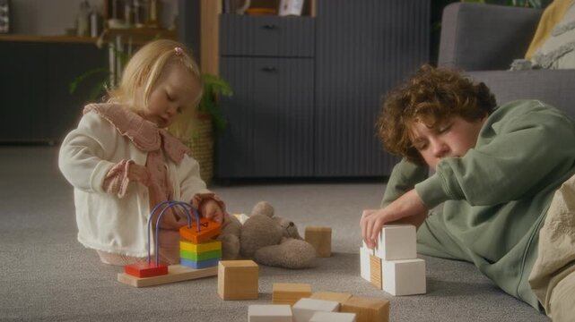 Side view shot of teenage brother and little sister playing together on floor with wooden toys, young boy babysitting child at home, copy space