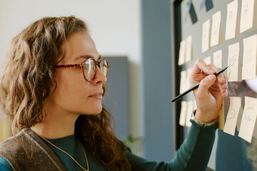 Caucasian young adult woman writing on sticky notes attached to glass wall, using pen while organizing tasks, wearing eyeglasses, focusing on project planning in modern workspace
