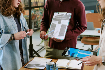 Young adult Caucasian woman, young adult Caucasian man, and young adult woman of color collaborating in office, holding clipboard and digital tablet, discussing documents together