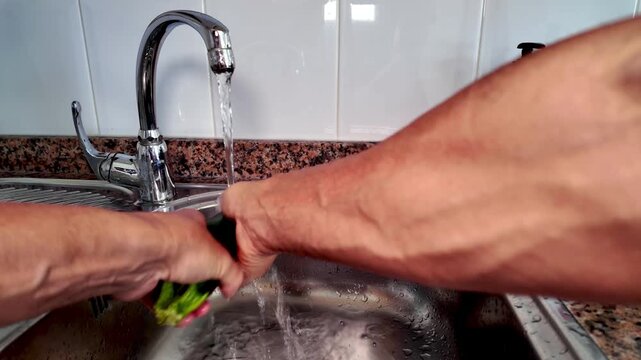 Man's hands washing a fresh green zucchini under running water in a stainless steel kitchen sink, preparing a healthy meal with fresh vegetables as part of a Mediterranean diet and Italian cuisine