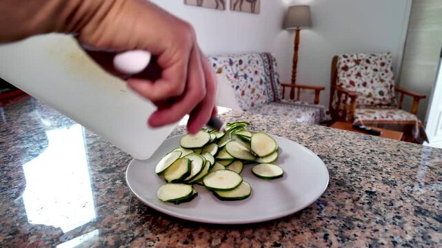 Cook's hands using a knife to move sliced fresh zucchini from a cutting board onto a white plate on a kitchen counter, a typical ingredient for a healthy Mediterranean diet at home
