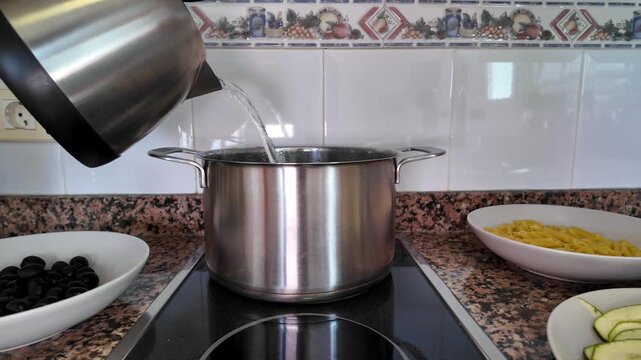 Person pouring hot water from an electric kettle into a stainless steel pot on an induction hob, preparing to cook a pasta dish with zucchini and olives, representing the Mediterranean diet