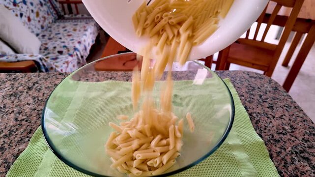 Preparing a typical Italian pasta dish, a staple of the healthy Mediterranean diet and a UNESCO intangible cultural heritage, by pouring cooked penne from a colander into a glass serving bowl