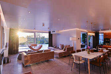 Fototapeta premium Modern luxury living room interior at sunset. Open-plan space featuring beige sofas, marble dining table, large glass windows with sun flare, and view of a patio. Stylish contemporary home design