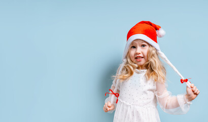 Cheerful little child girl in a white dress and Santa hat on a blue background, the concept of new year and Christmas, space for text on the banner
