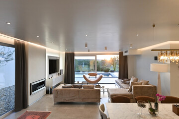 Fototapeta premium Modern luxury living room interior at sunset. Open-plan space featuring beige sofas, marble dining table, large glass windows with sun flare, and view of a patio. Stylish contemporary home design