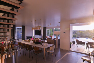 Fototapeta premium Modern luxury living room interior at sunset. Open-plan space featuring beige sofas, marble dining table, large glass windows with sun flare, and view of a patio. Stylish contemporary home design