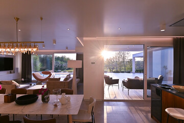 Fototapeta premium Modern luxury living room interior at sunset. Open-plan space featuring beige sofas, marble dining table, large glass windows with sun flare, and view of a patio. Stylish contemporary home design