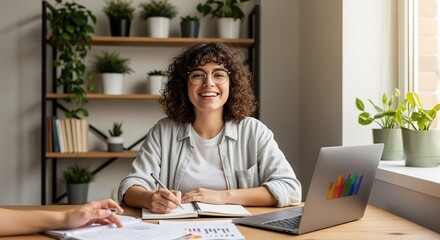 A young woman with curly hair smiles while working at a desk. She writes in a notebook with a laptop and papers nearby. Plants on shelves and a window create a bright, productive workspace.