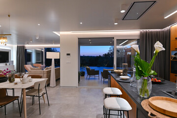 Modern luxury living room interior at sunset. Open-plan space featuring beige sofas, marble dining table, large glass windows with sun flare, and view of a patio. Stylish contemporary home design
