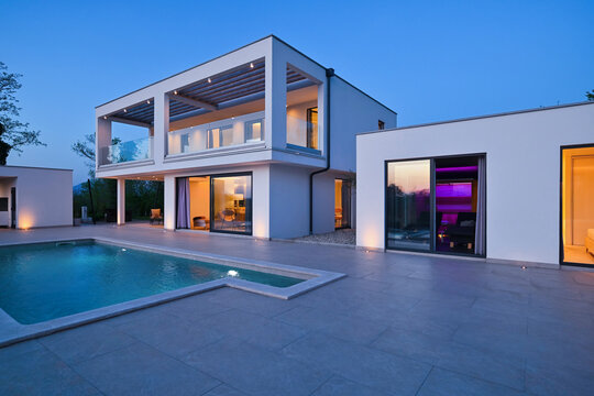 Exterior view of a modern luxury house with a swimming pool at twilight. Illuminated two-story white villa with large glass windows, patio, terrace, and glowing blue water