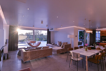Fototapeta premium Modern luxury living room interior at sunset. Open-plan space featuring beige sofas, marble dining table, large glass windows with sun flare, and view of a patio. Stylish contemporary home design