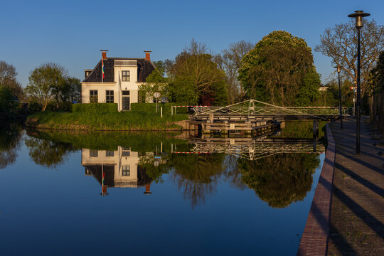 Perfect reflection of a white house and drawbridge in the calm canal in Bedum, Groningen. Bright spring morning and sun shadows on the sidewalk