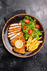 Roast viennese schnitzel steak, breaded meat with salad and french fry on a plate. black background. top view