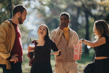 A diverse group of friends chat and snack in a sunny park, sharing popcorn and drinks while smiling and relaxing outdoors. The scene captures casual social moments and warm, friendly energy.