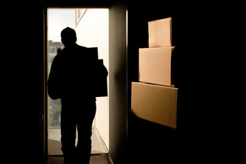 Returning unwanted gifts can be emotional experience as person leaves building carrying boxes, silhouetted in hallway with stacked cardboard packages
