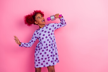 Young girl singing into a microphone against a pink backdrop in a purple polka dot dress