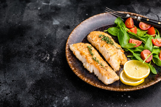Roast Cod fish fiilets, white fish meat on plate with salad. Black background. top view