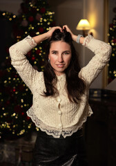 Woman in a Christmas decorated room poses in front of a tree, holding a red gift with ribbon and smiling. She places a large bow on the Christmas tree, festive interior, holiday mood.