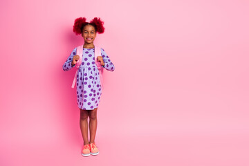 Young girl with pink backpack standing against pink background wearing a purple polka dot dress and bright sneakers