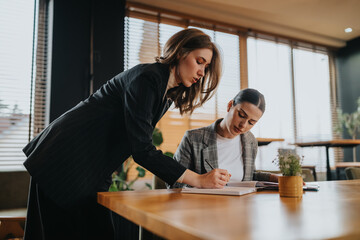 Two professionals work together in a modern office setting: one stands to write in a notebook while the other plans at the desk. Bright light, plants, and a calm, productive atmosphere.