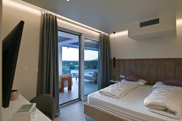 Fototapeta premium Bright, modern bedroom featuring a comfortable bed, sleek desk, and a large sliding glass door opening to a private balcony with stunning green landscape views, perfect for relaxation and serene