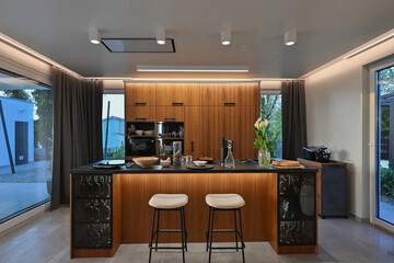 Modern luxury kitchen interior with wooden cabinetry, central island with bar stools, and ambient LED lighting. Stylish contemporary design with large windows and dark countertops at twilight