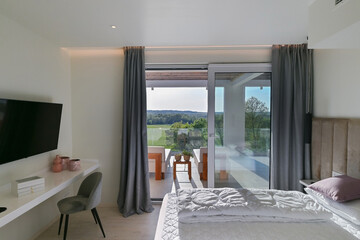 Obraz premium Bright, modern bedroom featuring a comfortable bed, sleek desk, and a large sliding glass door opening to a private balcony with stunning green landscape views, perfect for relaxation and serene