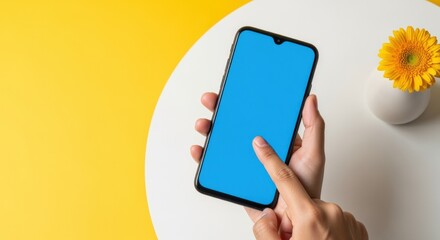 Hand Holding Smartphone with Blank Blue Screen on Yellow and White Table with Flower
