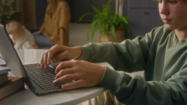 Rack focus shot of teenage boy using laptop at home with camera flowing from sister playing in background to hands typing on keyboard, copy space