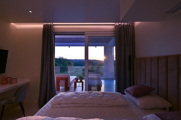 Obraz premium Bright, modern bedroom featuring a comfortable bed, sleek desk, and a large sliding glass door opening to a private balcony with stunning green landscape views, perfect for relaxation and serene