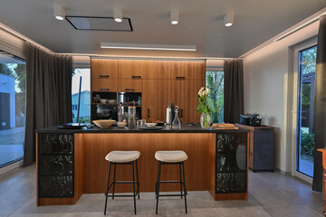 Modern luxury kitchen interior with wooden cabinetry, central island with bar stools, and ambient LED lighting. Stylish contemporary design with large windows and dark countertops at twilight