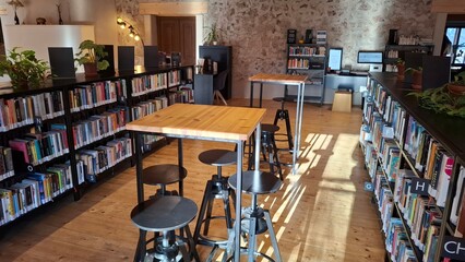 Rows of wooden tables and tall stools in a library with bookshelves and computers during daylight in a quiet space