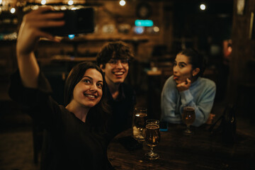 A group of teenage friends sits around a wooden table in a warm, dimly lit bar, capturing a cheerful selfie as drinks glitter in the foreground, sharing laughter and casual conversation.