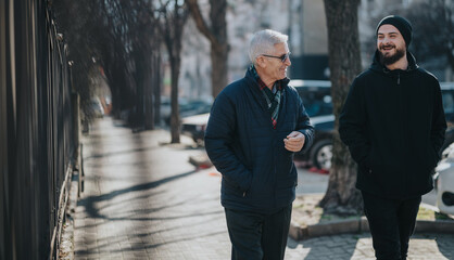 Men share a friendly conversation while strolling along a city sidewalk. The older man wears a blue...