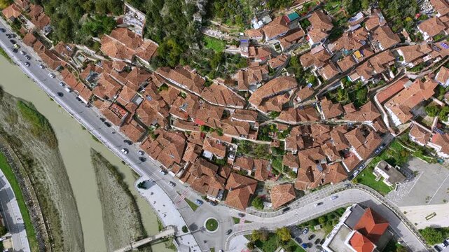 Aerial video of Berat featuring dense traditional rooftops, the riverside boulevard, a roundabout and the pedestrian bridge spanning the Osum River.