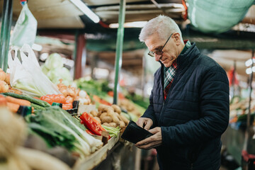 An elderly man in a dark jacket browses fresh produce at a vibrant market stall, checking his wallet for payment. Colorful vegetables, bags, and market ambiance surround him.