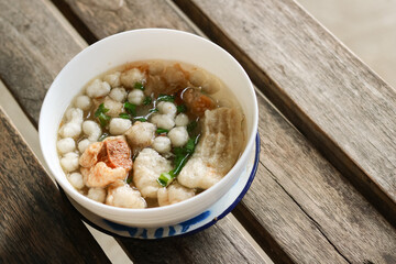 A portion of meatballs served in a white bowl containing clear broth, fried tofu, peanuts, fresh spring onions, and meatballs. The photo was taken from a high angle from a close distance with the bowl