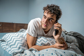 Young man having a conversation on his smartphone from bed, enjoying modern communication in morning at home