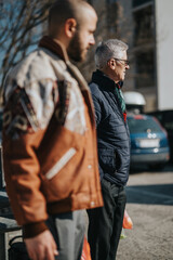 Obraz premium Two men stand on a sunny city street, one elderly man in a dark quilted jacket with scarf and glasses, the other in a brown jacket. They pause, observing the scene around them.