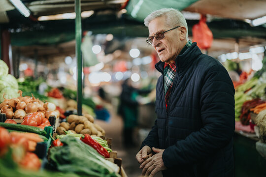 An elderly man in a dark jacket browses a colorful market stall lined with onions, peppers, and greens. The scene captures daily life, community, and the warm, bustling mood of a local market. - Powered by Adobe