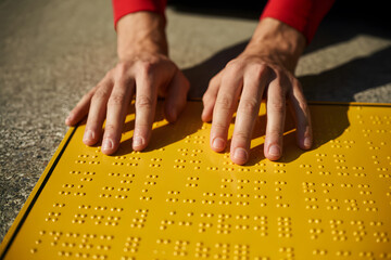 Close-up of hands in red sleeves gently touching a bright yellow tactile panel with raised black braille, set against a blurred, textured concrete background.