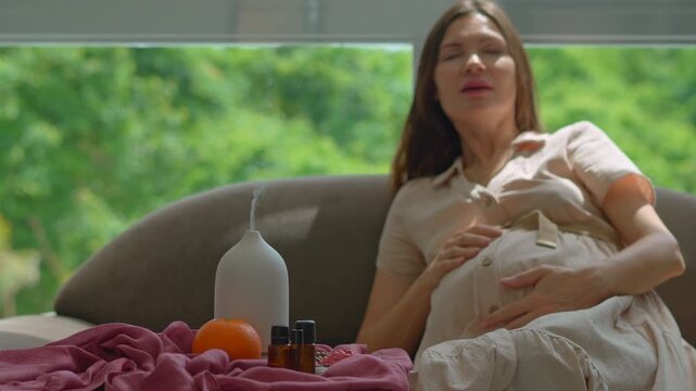 Pregnant woman over 40 sitting on a couch by a sunlit window enjoying scent and relaxation from a working electric aroma diffuser. Wellness, pregnancy care and relaxation lifestyle concept. Slowmotion
