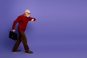 Elderly gentleman in red sweater with briefcase walking against purple background checking his watch