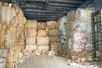 Large stacks of compressed paper blocks are piled high inside a dark industrial building. The paper appears to be waste material ready for recycling at the paper mill