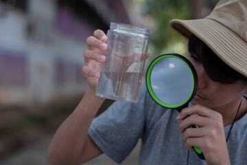A boy, geared up with a cream boonie hat and magnifying glass, passionately exploring tiny life forms contained within a clear plastic bottle. Igniting curiosity and the spirit of scientific discovery