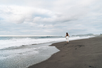 Girl Running Along the Gray Volcanic Santa Bárbara Beach by the Atlantic Ocean in a White Dress, Back to the Camera, São Miguel, Azores © Анико Мкртычян