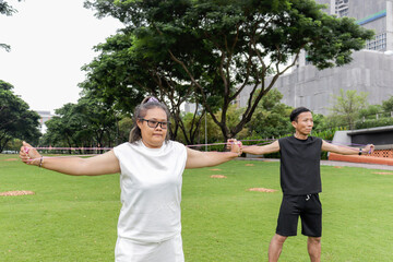 Plump middle-aged Asian woman and friend stretching body, doing exercise with resistance band with at park. Chubby female with buddy doing work out together outdoors. People with diversity real body.
