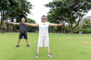 Plump middle-aged Asian woman and friend stretching body, doing exercise with resistance band with at park. Chubby female with buddy doing work out together outdoors. People with diversity real body.